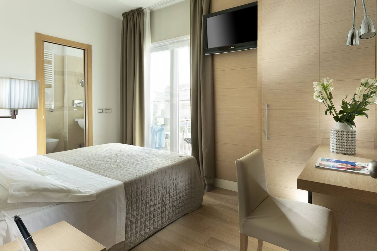 Una habitación de hotel con cama, escritorio, televisor y ventana con cortinas.