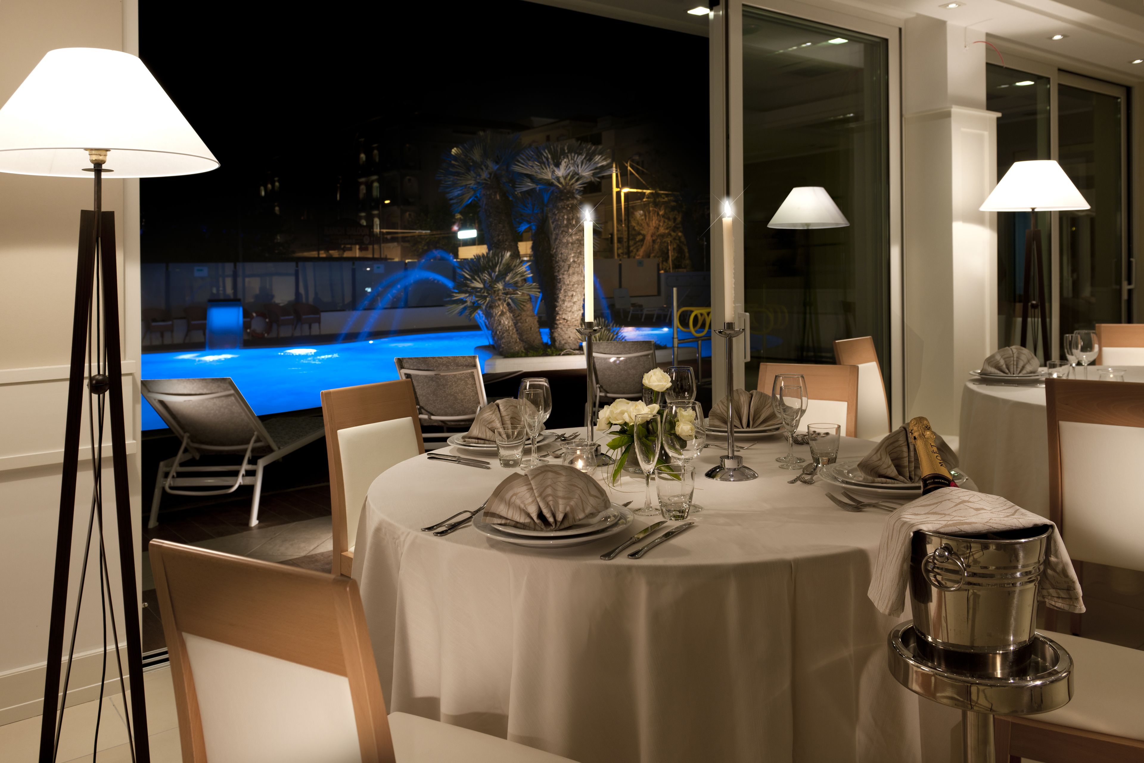 A dining area with a table set for a meal, a champagne bottle, and a view of a pool at night.