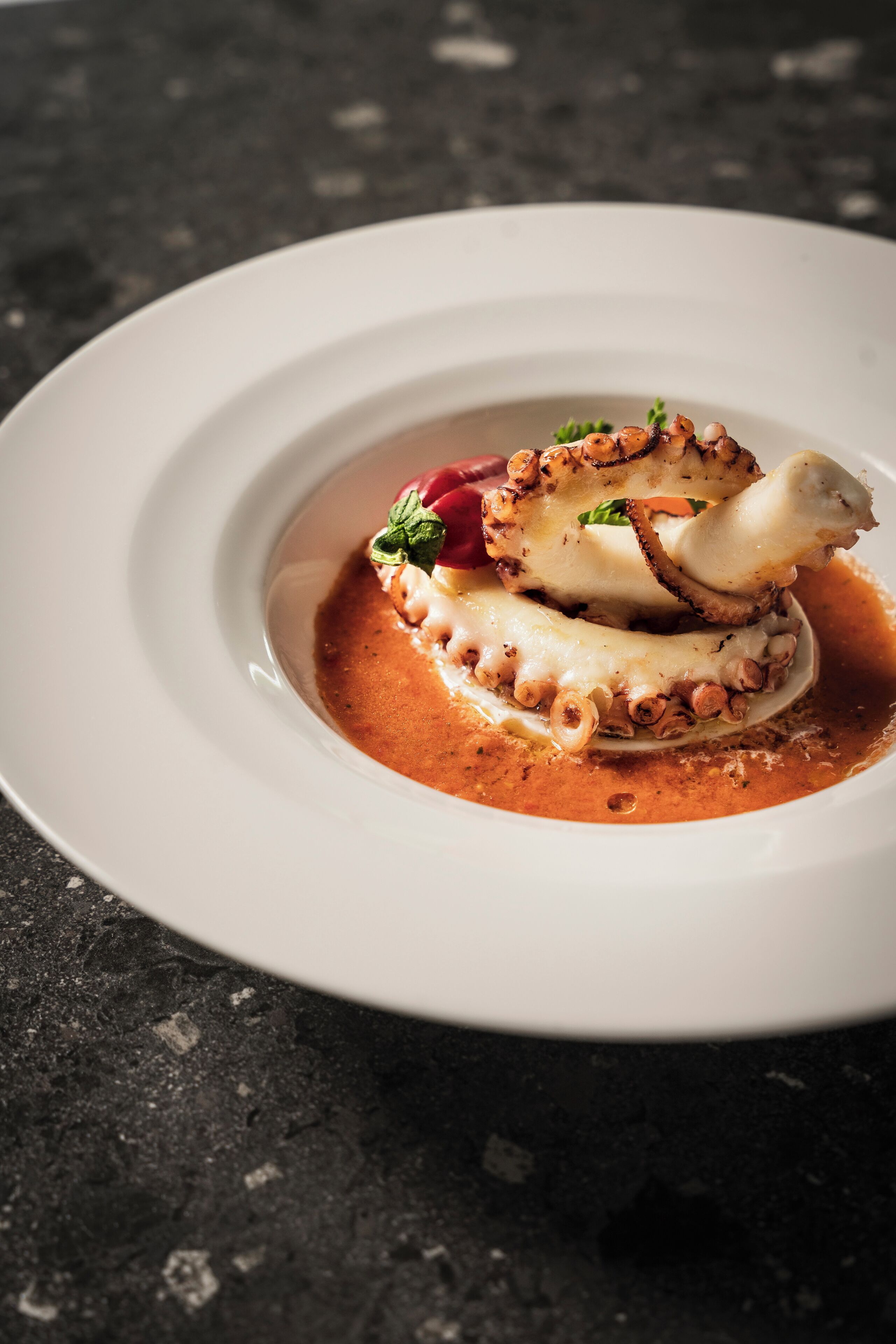 Une assiette blanche contenant un plat de fruits de mer, peut-être des poulpes, agrémenté d’herbes et d’un fruit rouge.