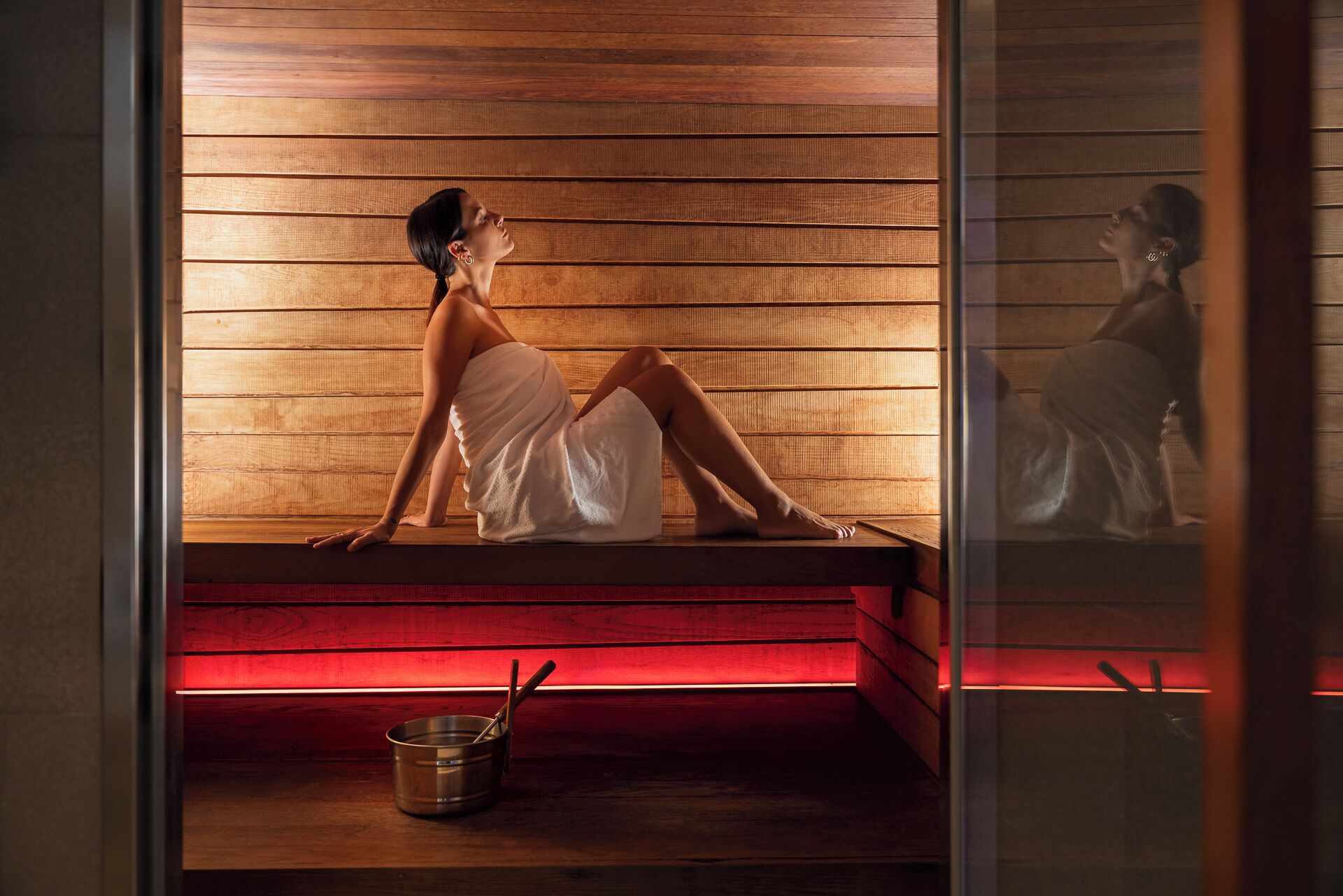 Une personne assise dans un sauna, avec un mur en bois et un banc éclairé de lumière rouge.