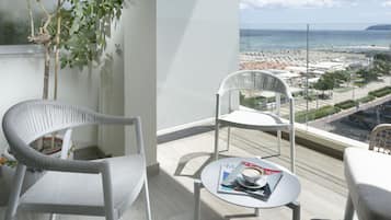 Un balcon avec deux chaises blanches, une petite table sur laquelle se trouvent un magazine et une tasse, et une vue sur une plage et le paysage urbain.