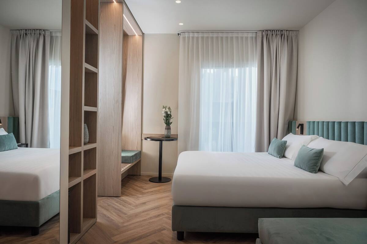 Habitación de hotel con dos camas, un armario de madera, una mesita con un florero y cortinas traslúcidas.