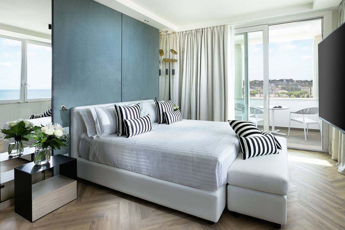 Un dormitorio moderno con una cama grande, una mesita de noche con un jarrón de flores y vistas a la ciudad por la ventana.