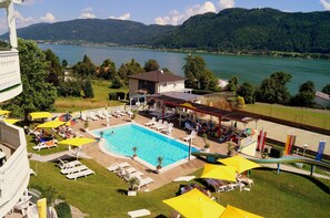 Une piscine dotée de chaises longues et de parasols, entourée d’un bâtiment avec une zone couverte et offrant une vue sur un lac et des montagnes.
