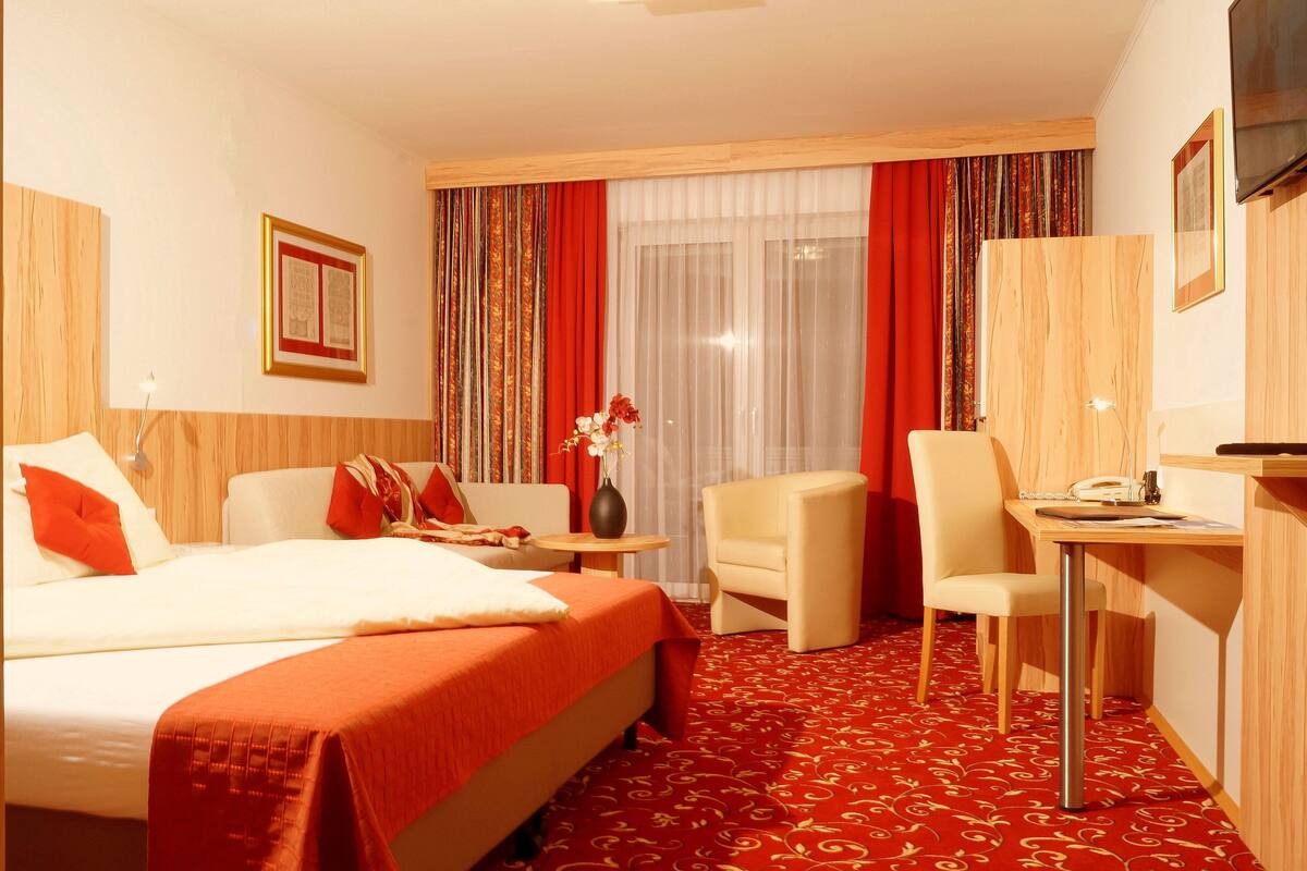 A hotel room with a large bed, a desk, a chair, and a small table with a vase of flowers.