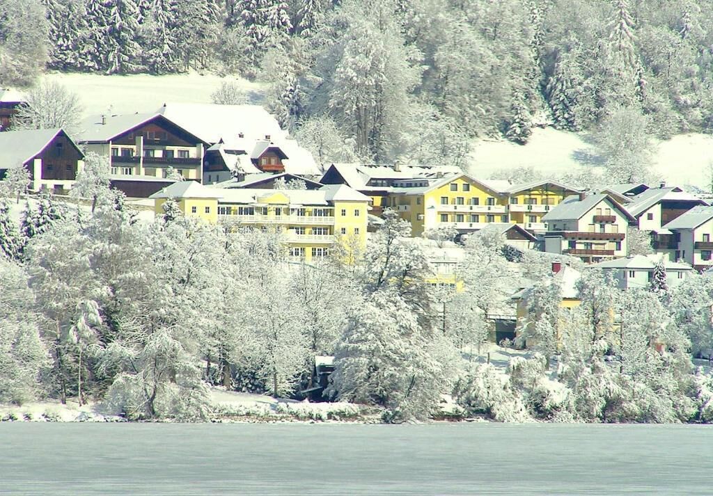 A cluster of multi-story buildings with balconies, situated by a body of water, surrounded by snow-covered trees.