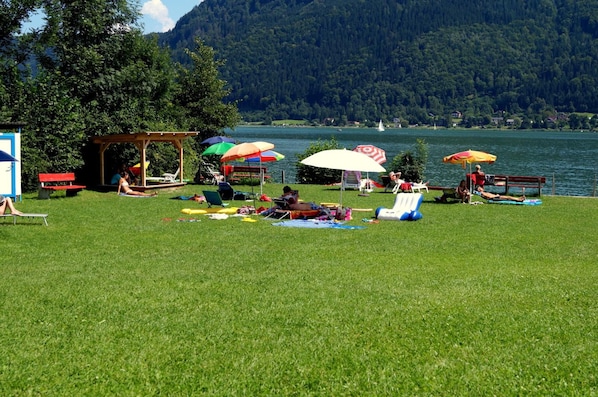 A grassy area with picnic tables and umbrellas near a lake, surrounded by trees and hills.