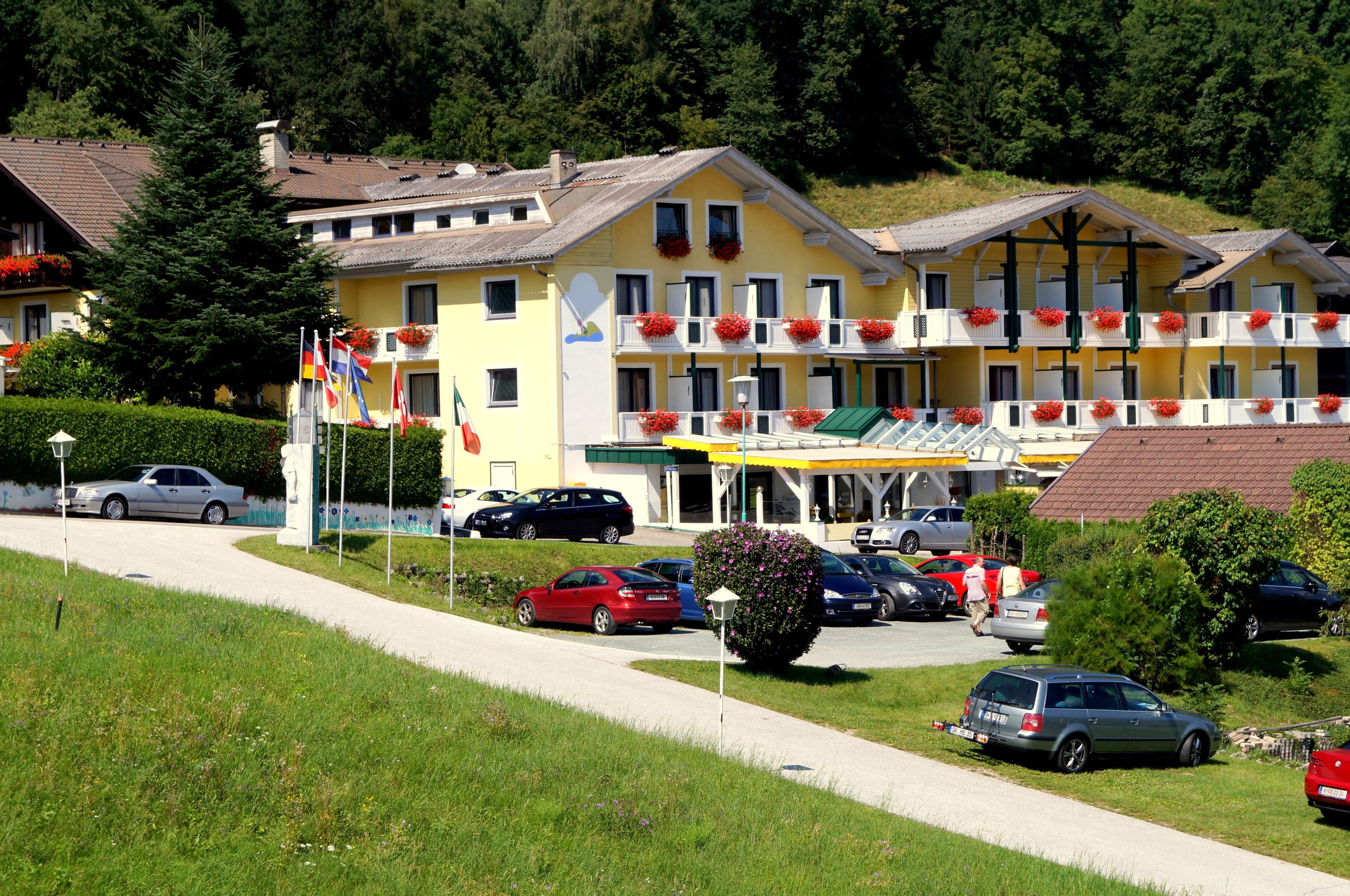 Ein mehrstöckiges Hotel mit Balkonen, die mit roten Blumen geschmückt sind, einem Parkplatz und einer Hanglage im Hintergrund.