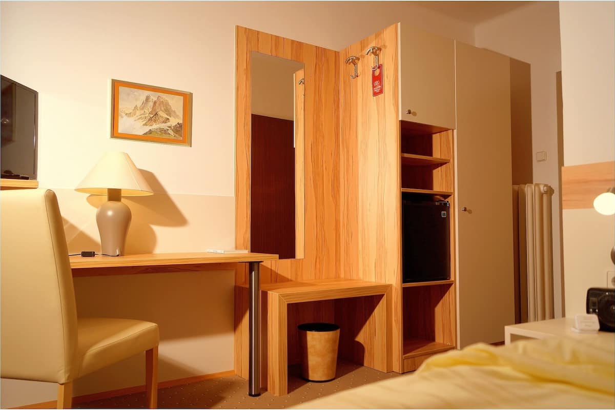 A hotel room with a wooden desk, chair, and a bed with a yellow blanket. There is a TV on the left and a painting on the wall.