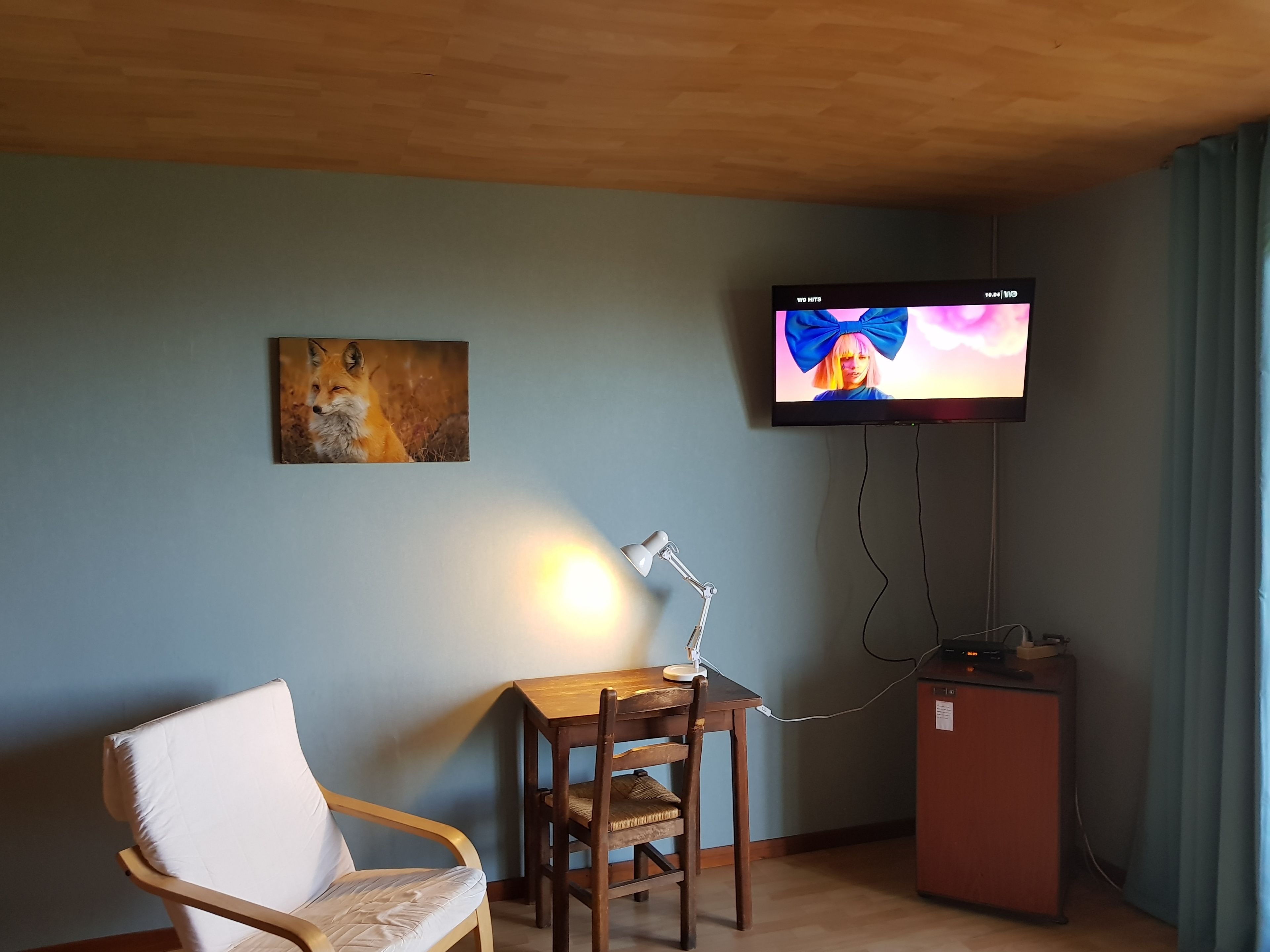Une pièce avec un plafond en bois, un téléviseur fixé au mur, un bureau avec une lampe, une chaise et un tableau accroché au mur.