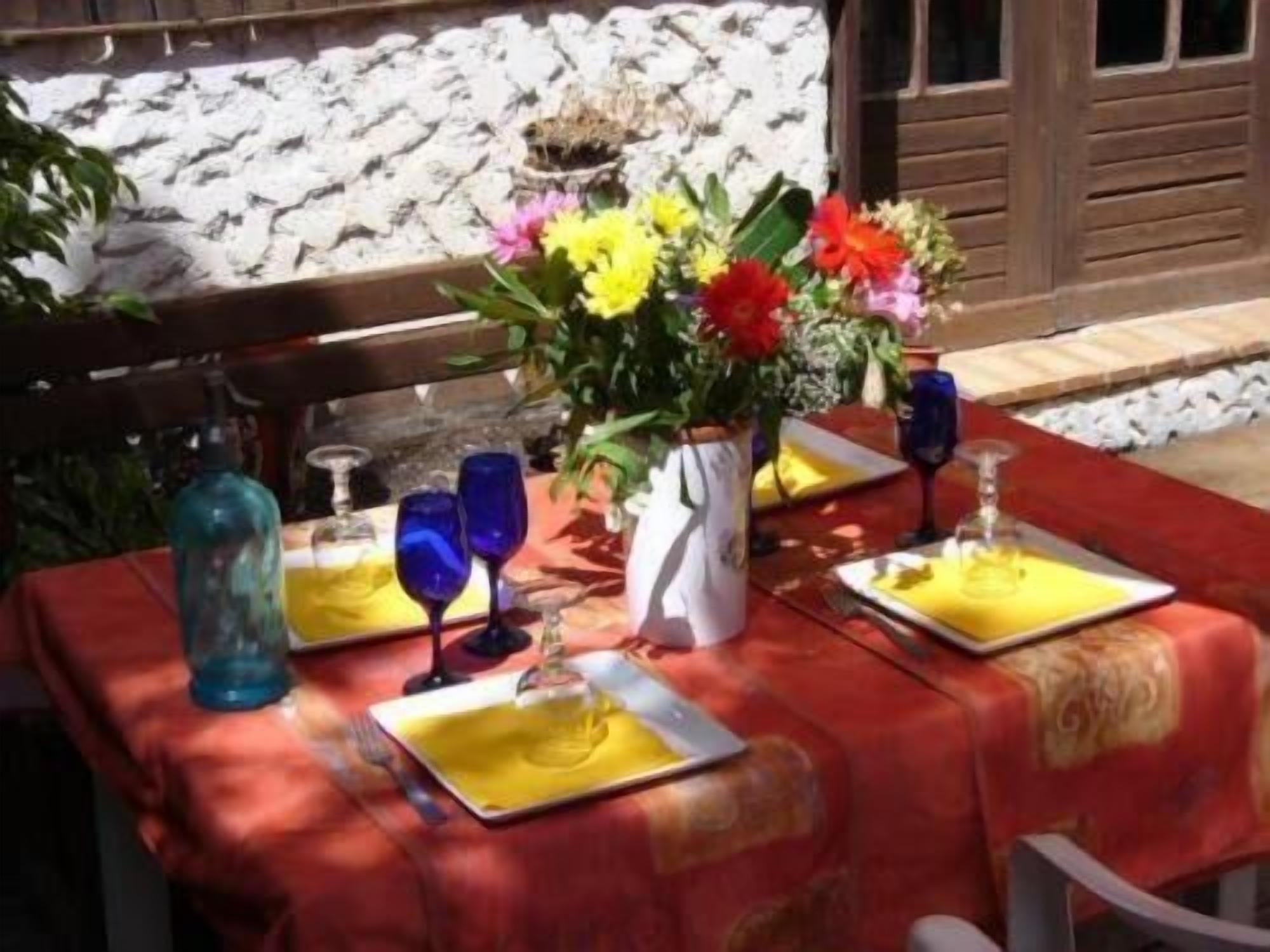 A table set outdoors with a red tablecloth, yellow napkins, blue glassware, and a vase with flowers.