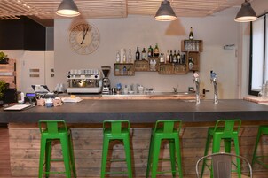 Angolo bar con un bancone in legno, sgabelli verdi, una macchina da caffè e scaffali con bottiglie e bicchieri.