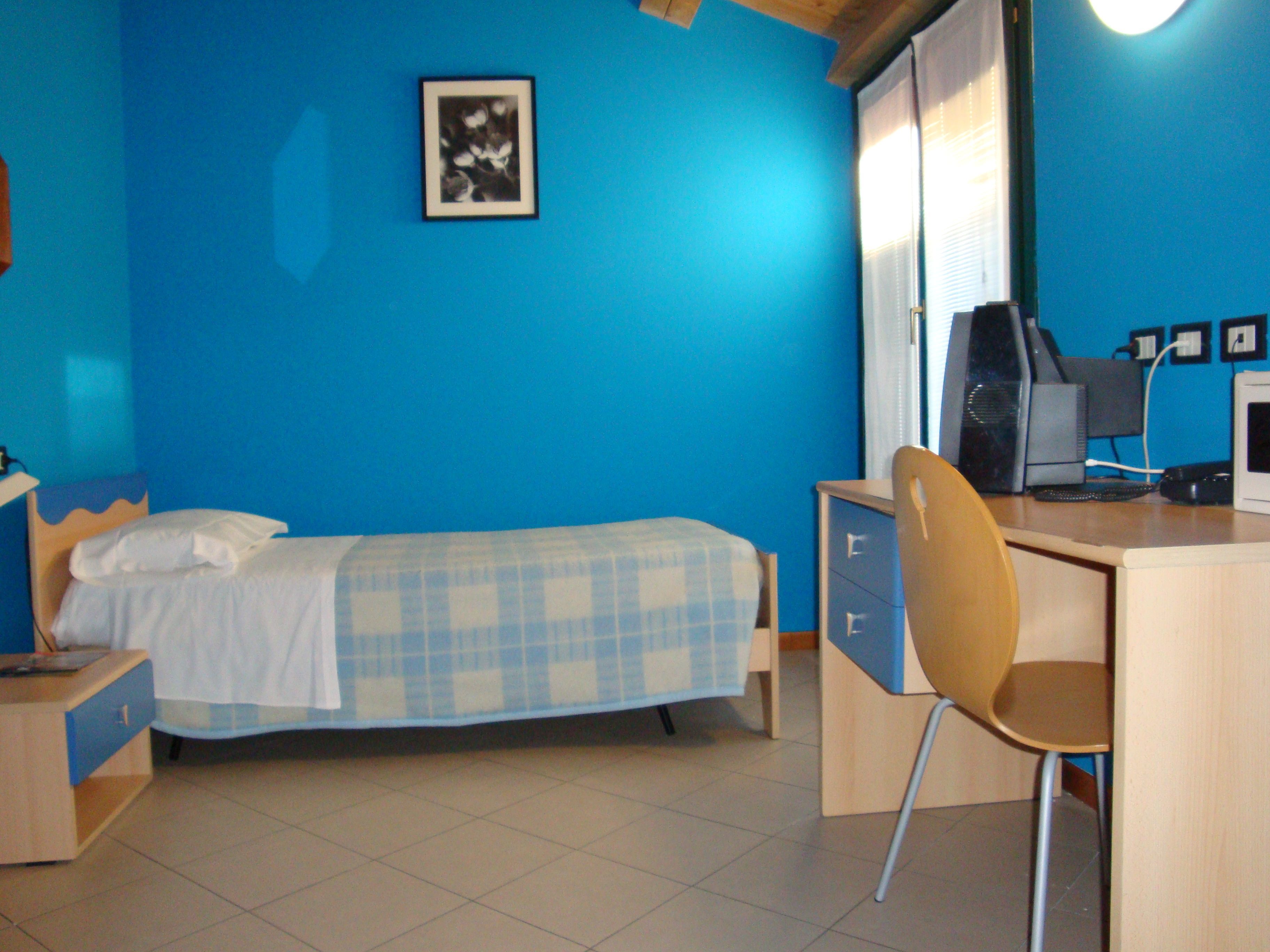 Una habitación con cama, escritorio, silla, computadora y un cuadro en la pared.