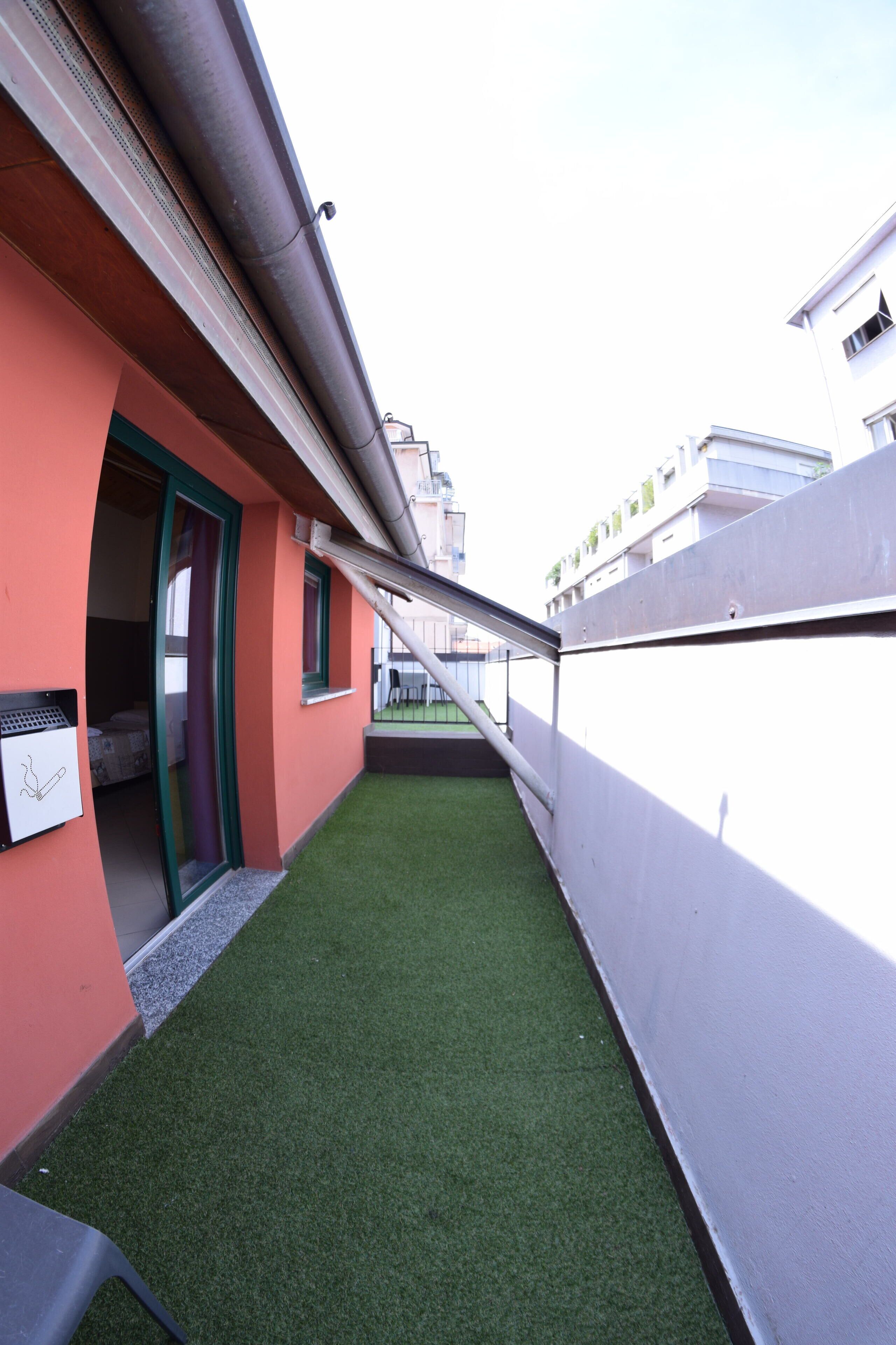 Un estrecho pasillo exterior con césped artificial verde, una pared blanca y un edificio rojo con puerta y ventana.