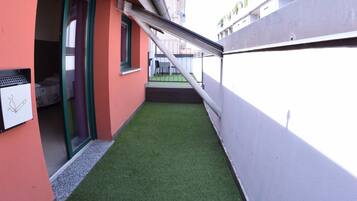 Un estrecho pasillo exterior con césped artificial verde, una pared blanca y un edificio rojo con puerta y ventana.