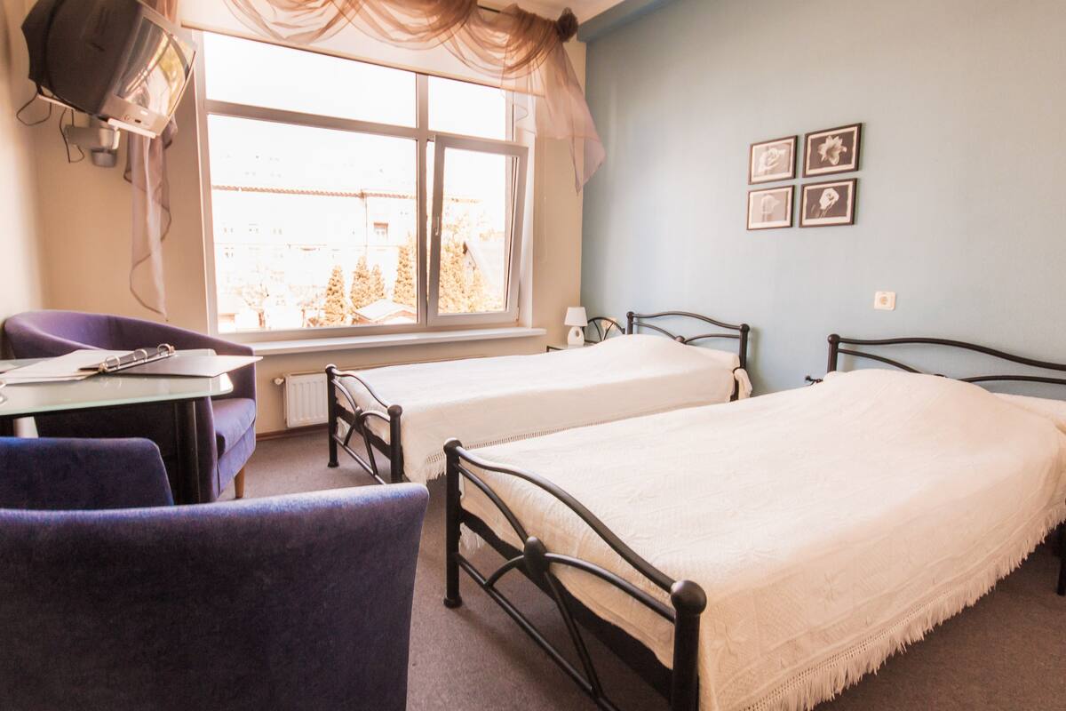 Una habitación con dos camas individuales, un escritorio con silla, una ventana con cortinas y un cuadro con marcos.