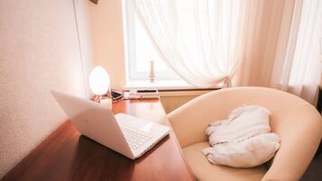 A laptop on a wooden desk, a chair with a white cushion, a mirror, and a window with sheer curtains.