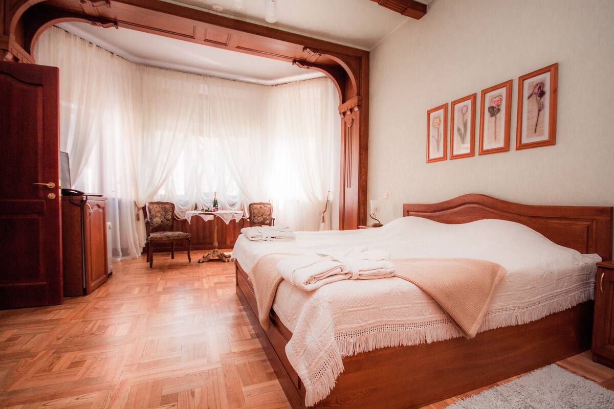 Un dormitorio con una cama de madera, una silla, un escritorio y un ventanal con cortinas translúcidas.
