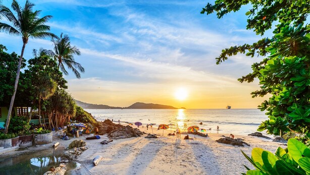 A beach with a sunset, palm trees, and umbrellas. - Silver Resortel (Patong)