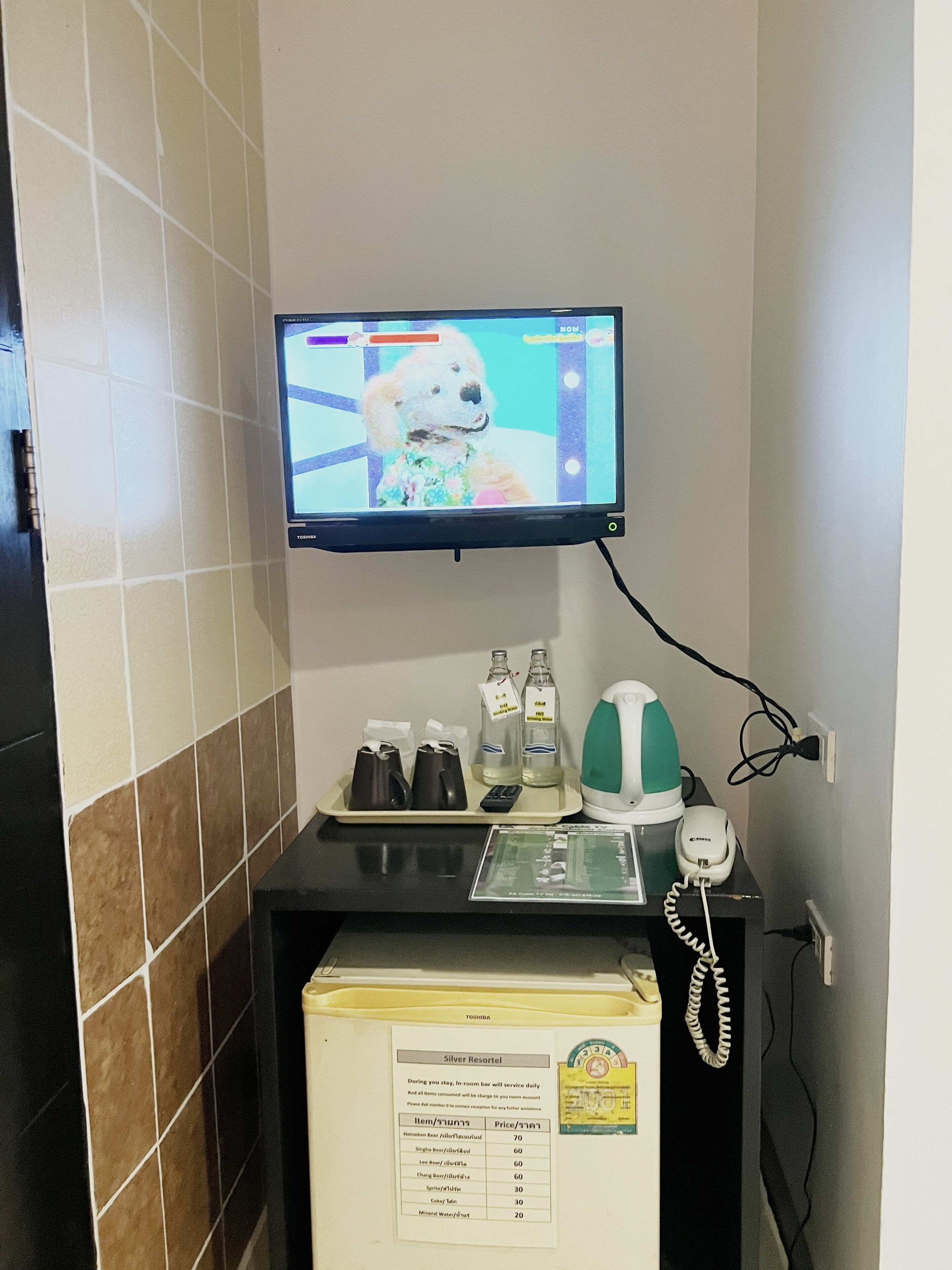 Hotellihuone, jossa on televisio, jossa näkyy kuva koirasta, minibaari, puhelin sekä tarjotinalusta pulloineen ja kaukosäädin.