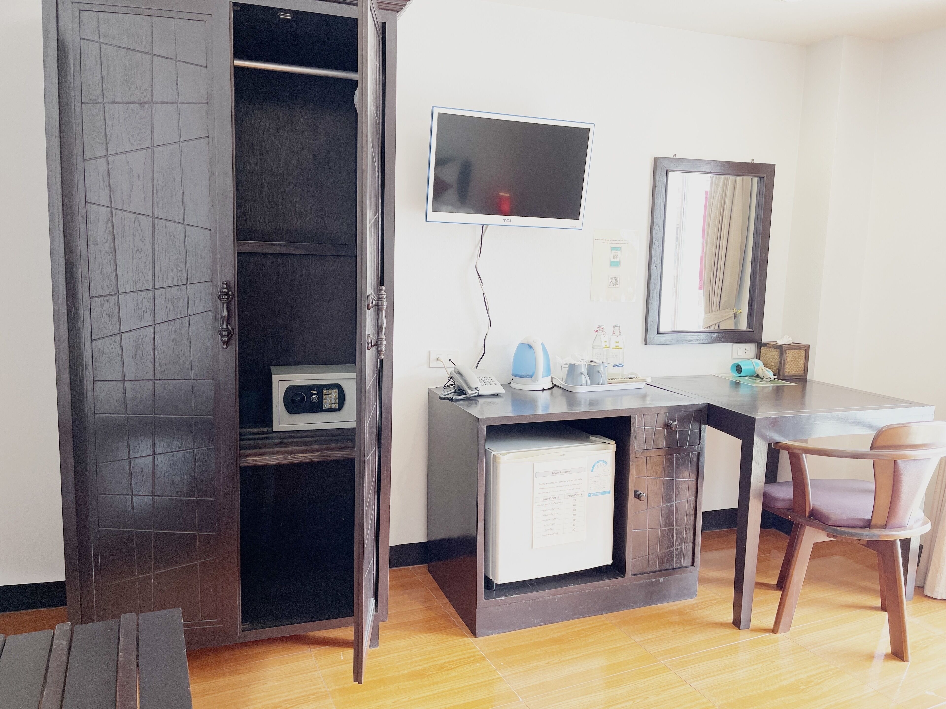 Una camera d'albergo con pavimento in legno, una scrivania con computer, una sedia, una cassaforte, una televisione e uno specchio.