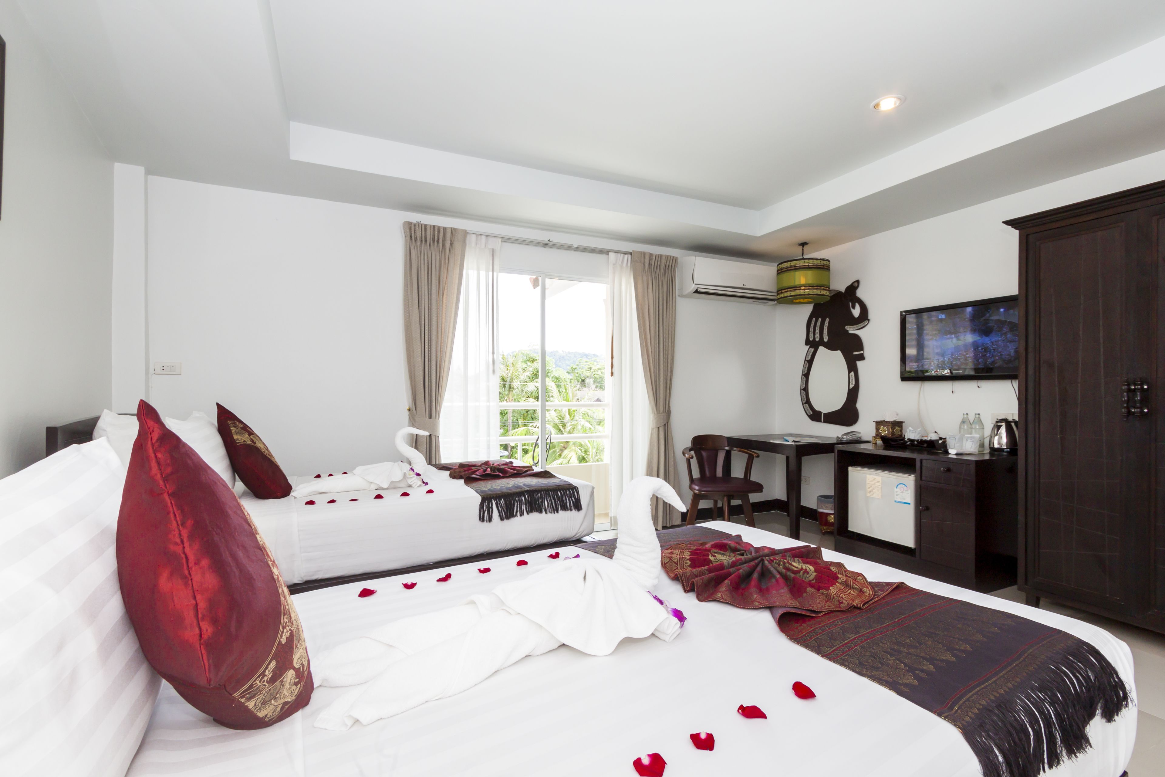A hotel room with two beds, a desk, a television, and a view of greenery outside.
