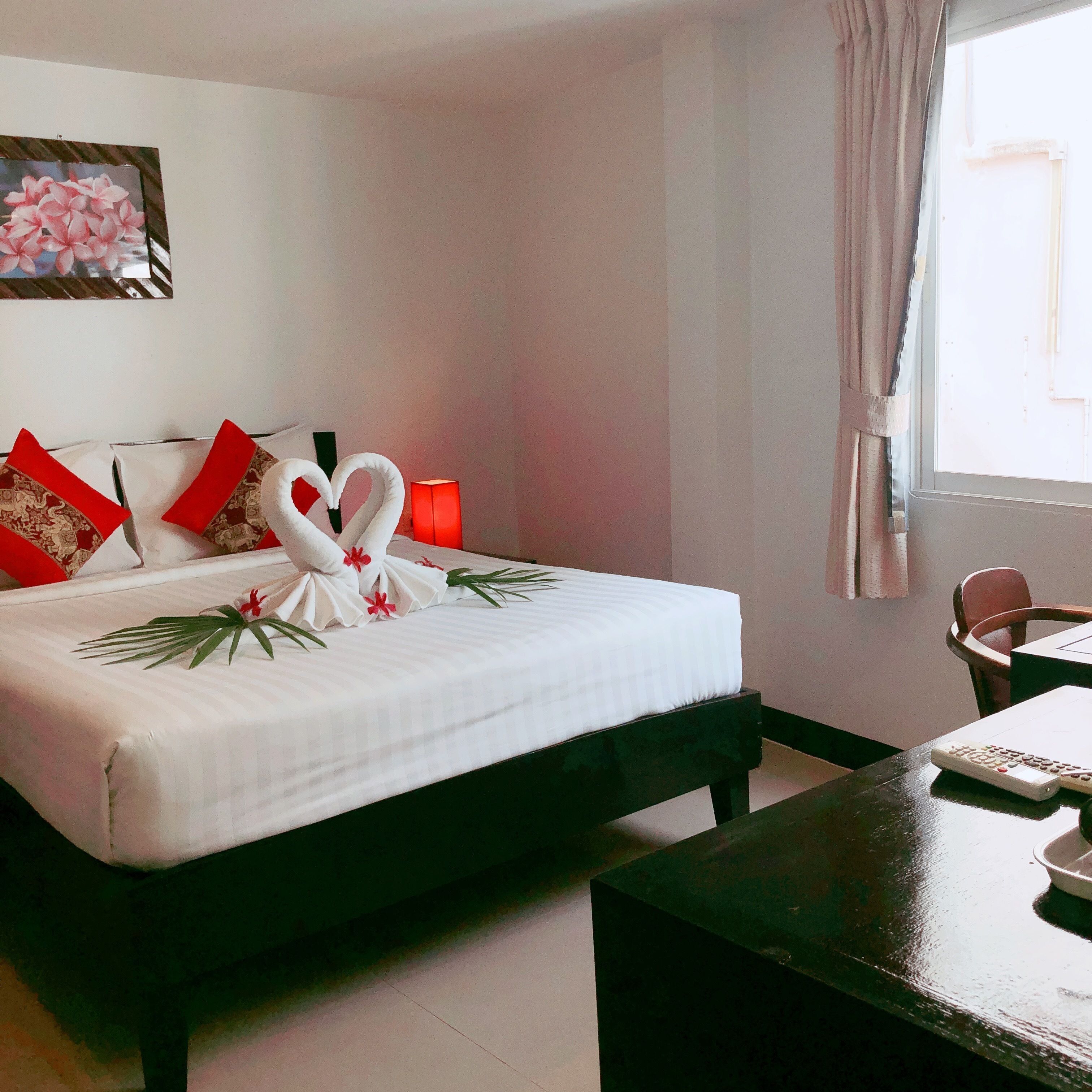 Una cama bien hecha con una colcha blanca, almohadas rojas y una decoración de toalla con forma de corazón. Hay una lámpara al lado de la cama y una mesita con un control remoto.