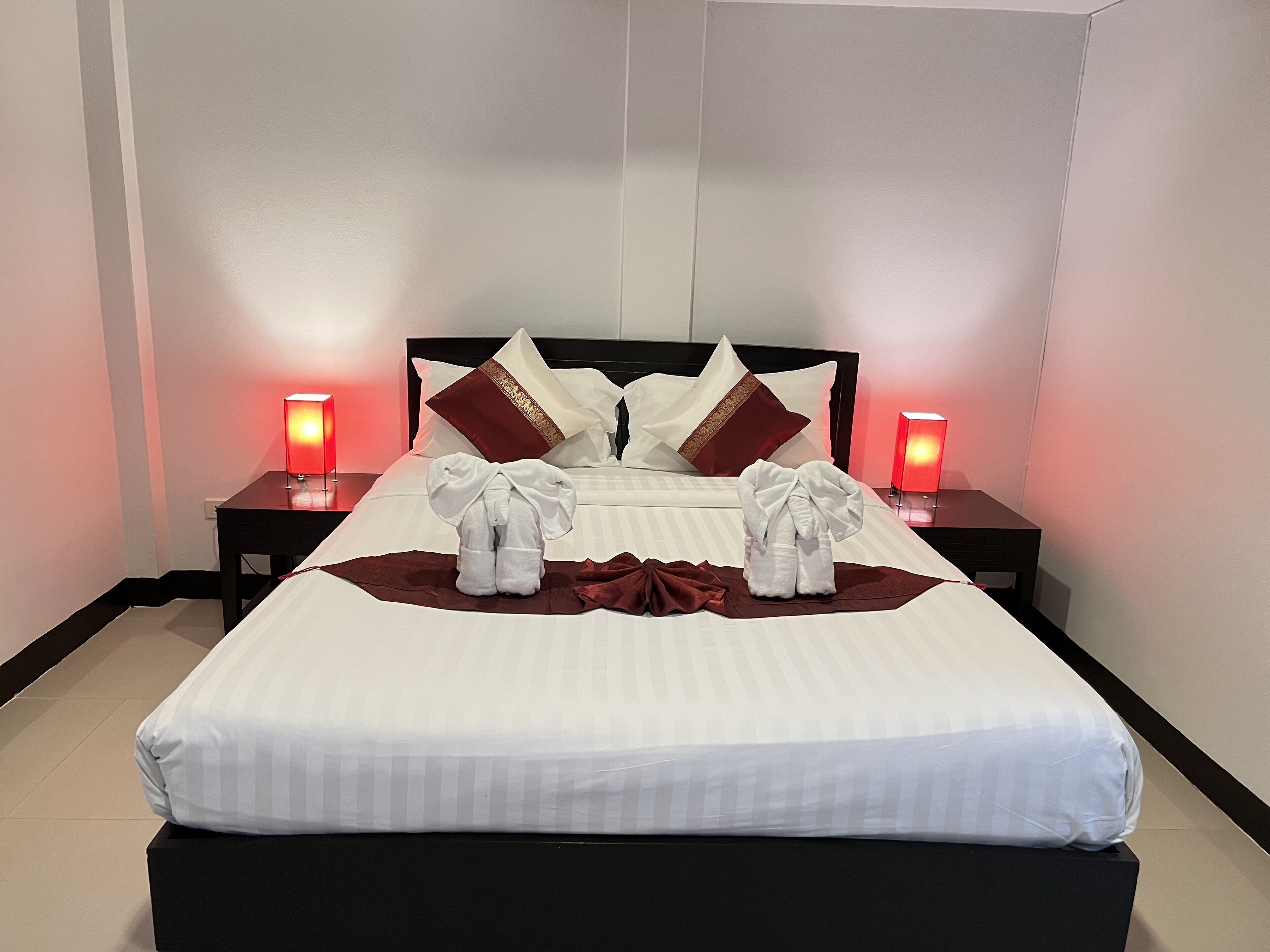 A neatly made bed with white linens and two towels arranged on a brown cloth in the center. There are two bedside tables with red lamps on either side of the bed.