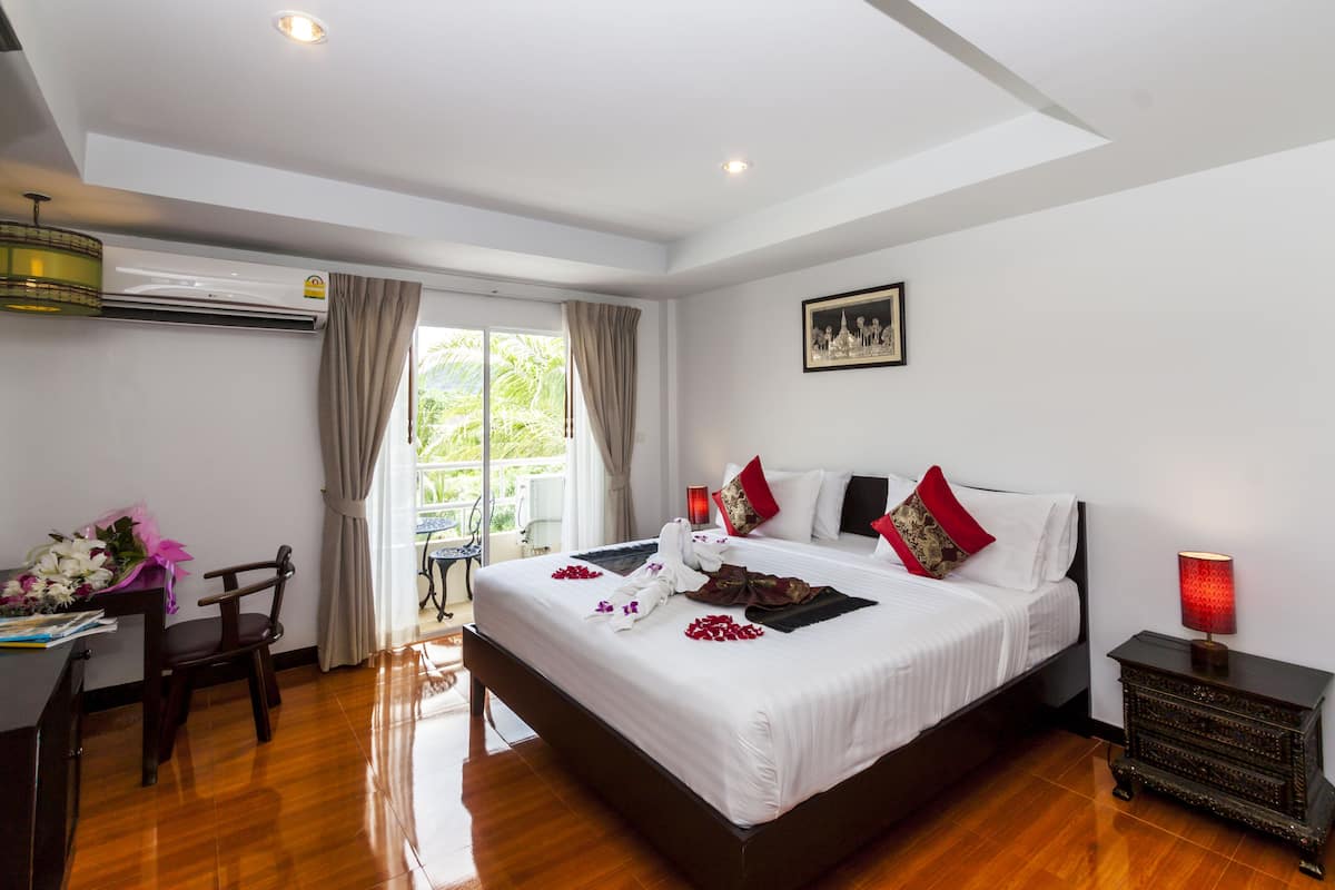 A hotel room with a large bed, a desk, a chair, an air conditioning unit, a window with curtains, and a nightstand with a lamp.
