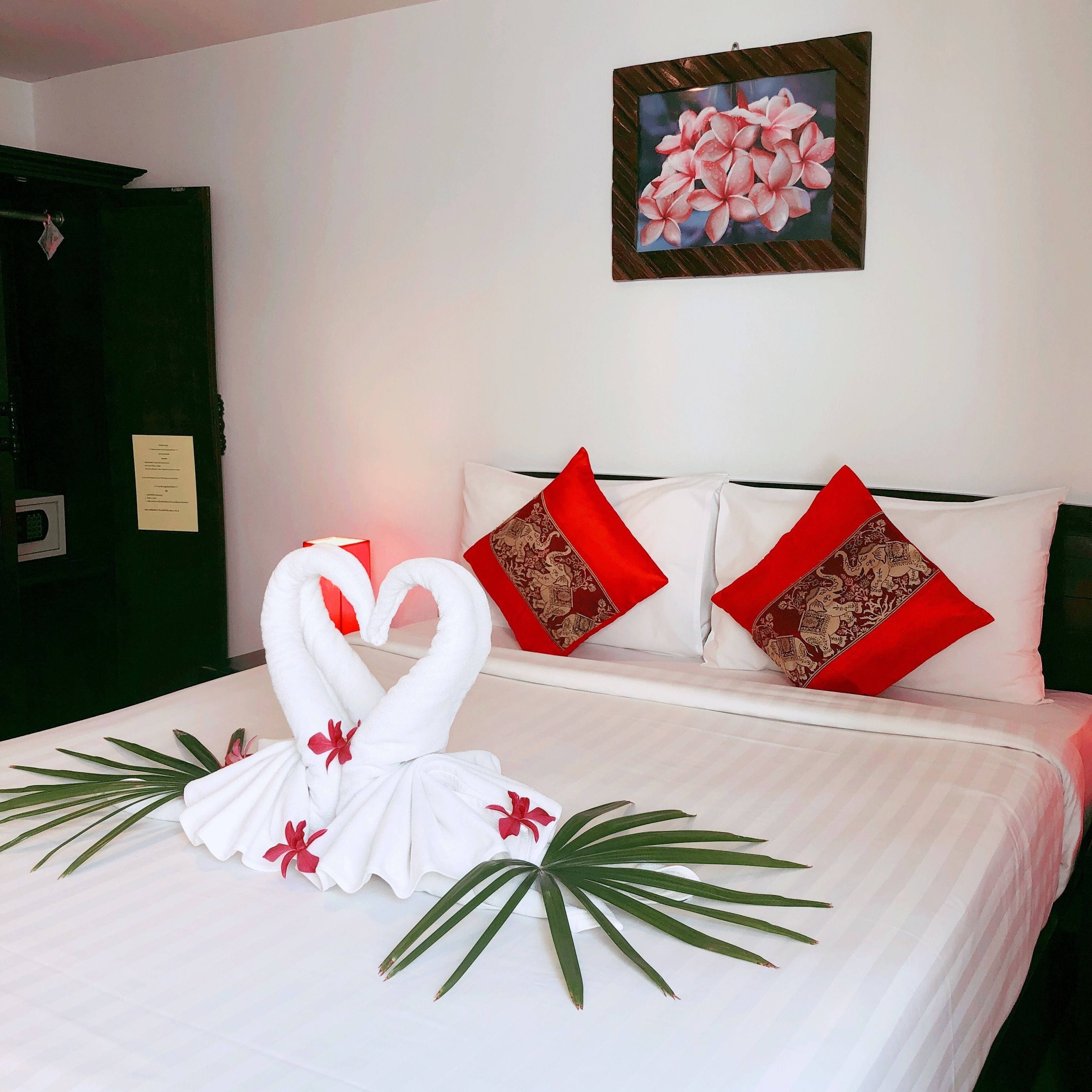 Un lit parfaitement fait, agrémenté d’une décoration centrale en forme de cygne réalisée avec une serviette, de coussins rouges et d’un tableau floral accroché au mur.
