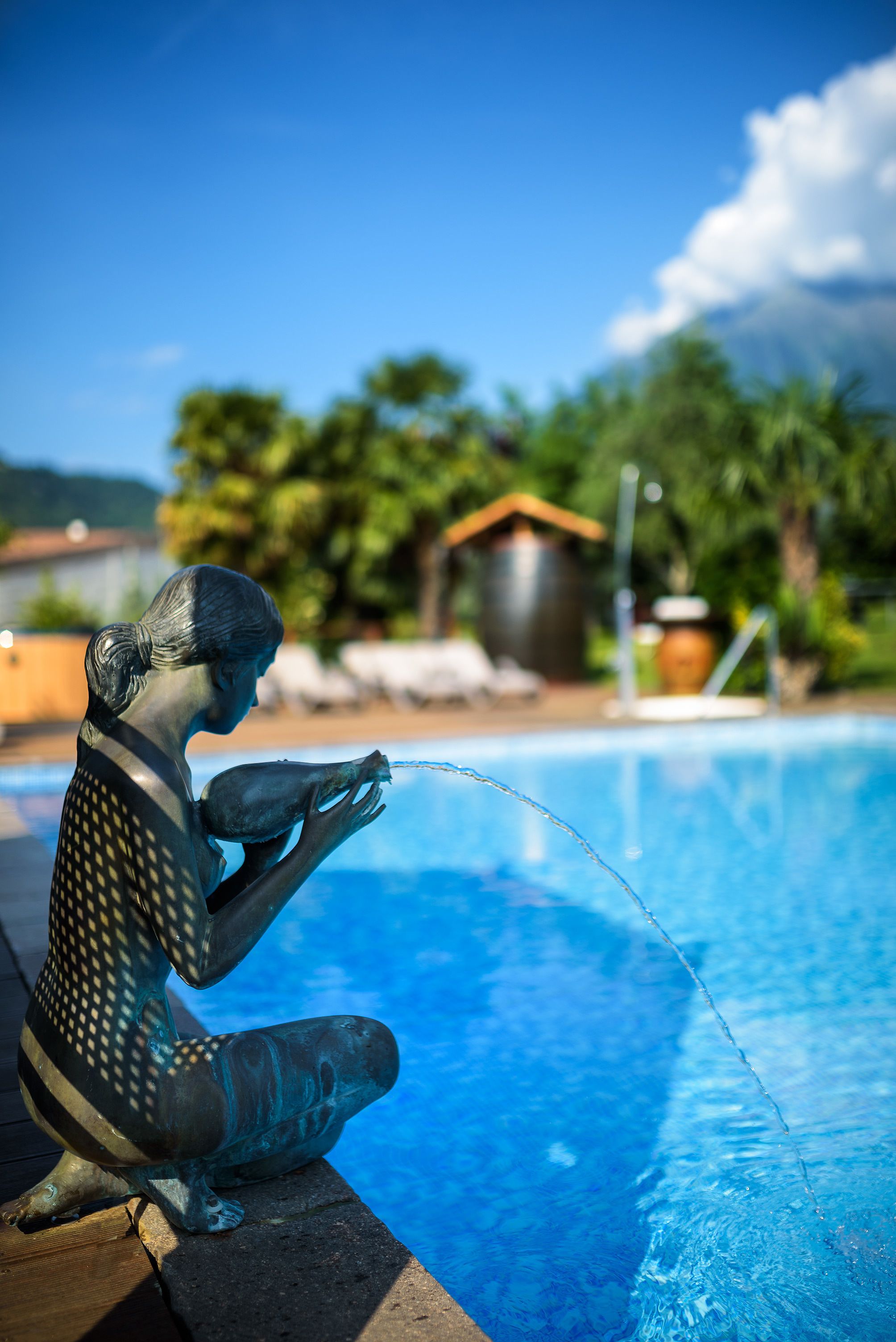 A statue of a woman by a pool with a clear blue sky and greenery in the background.