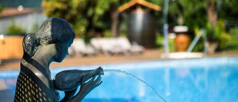 A statue of a woman by a pool with a clear blue sky and greenery in the background.