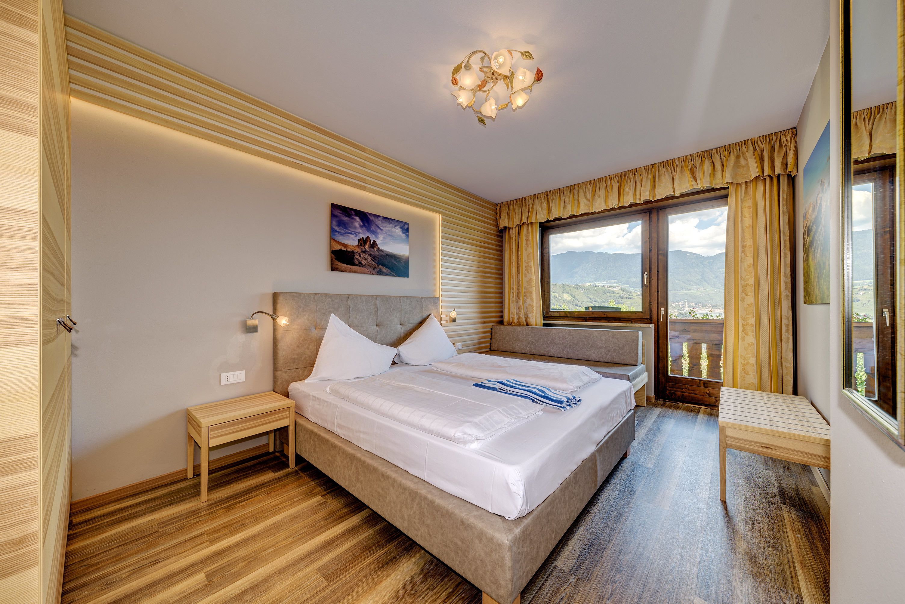 A bedroom with a large bed, wooden floor, a painting on the wall, and a view through the window.
