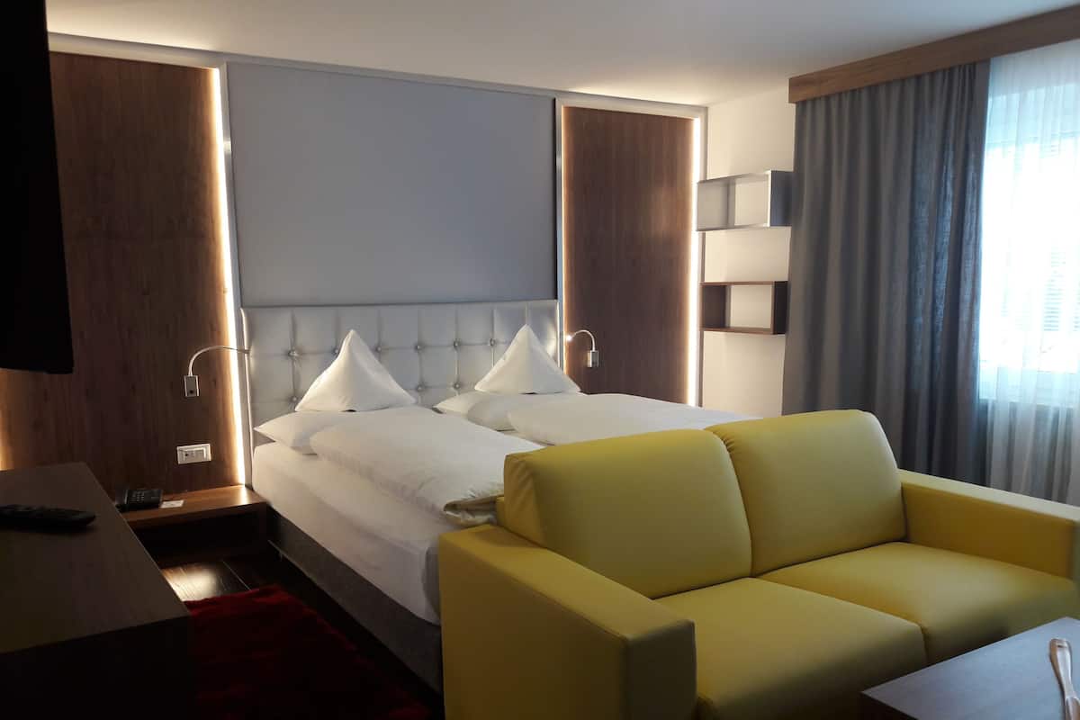 Quarto de hotel com cama, sofá amarelo, escrivaninha com telefone e televisão na parede.