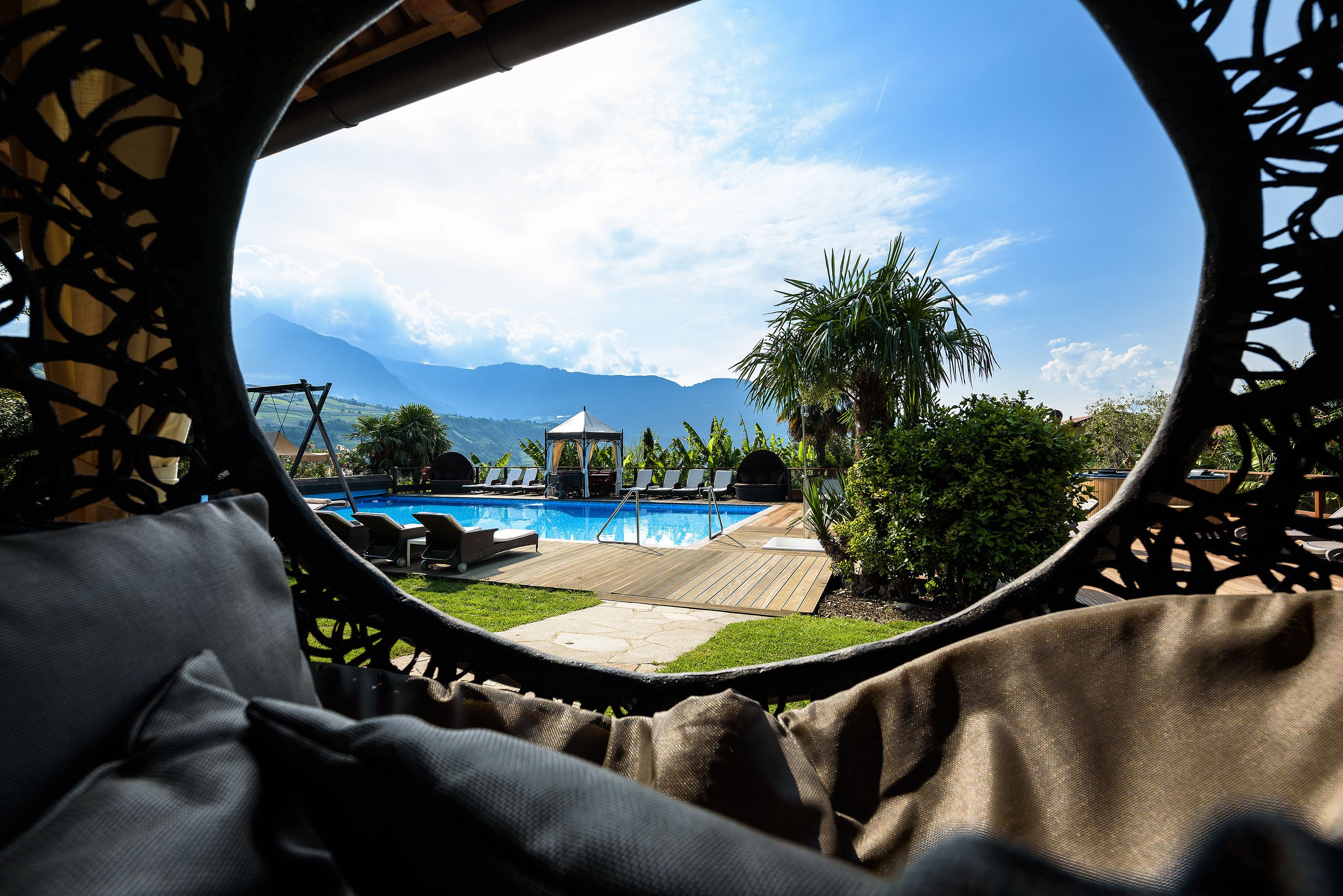 A view through a circular window of a pool area with lounge chairs, a gazebo, and mountains in the background.
