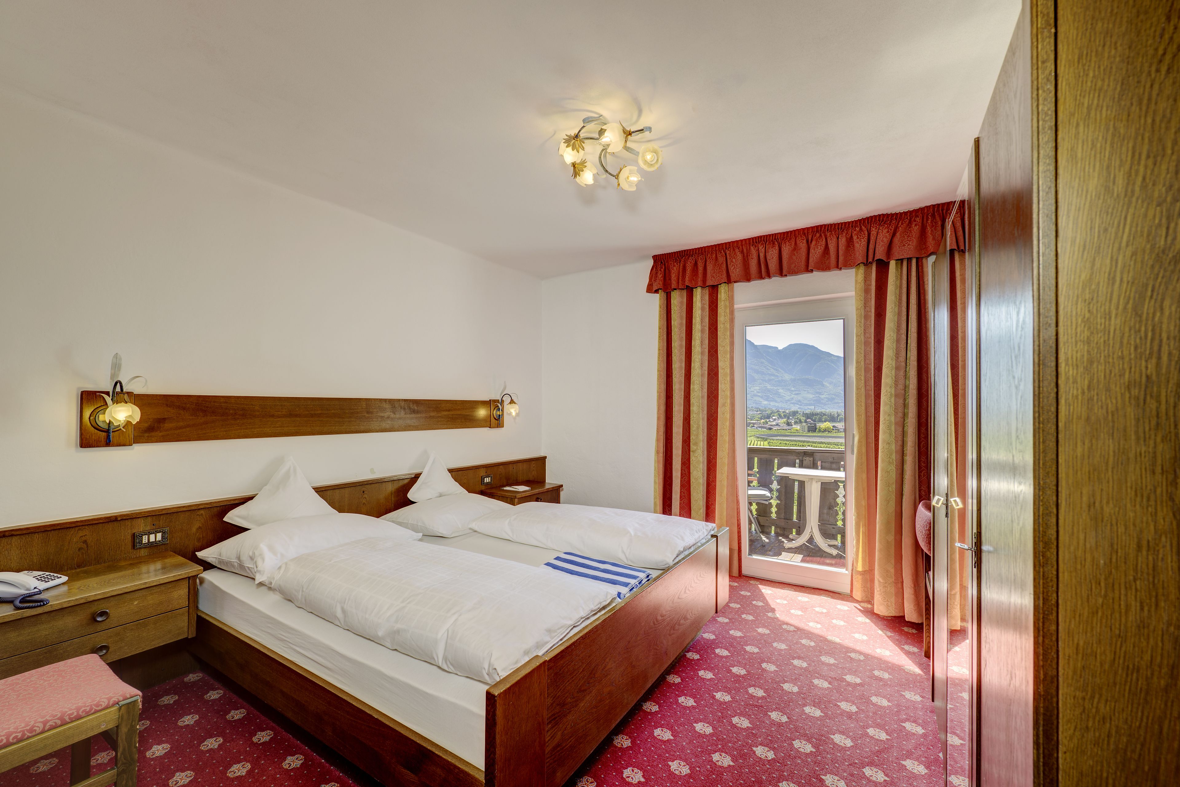 Habitación de hotel con cama de madera, mesita de noche, escritorio y silla. Tiene un balcón con vista a las montañas.