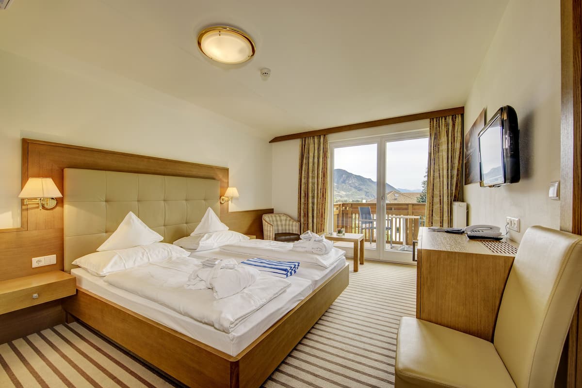 Quarto de hotel com uma cama grande, uma escrivaninha com televisão de tela plana, uma cadeira e varanda com vista para as montanhas.