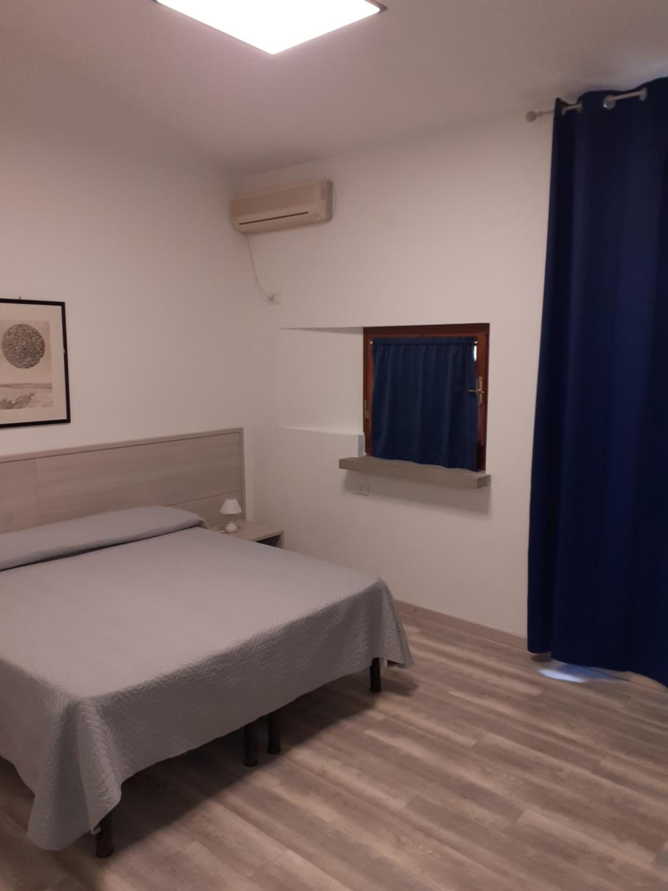Un dormitorio con una cama, una mesita de noche, un aire acondicionado instalado en la pared, una ventana con cortina y un cuadro en la pared.