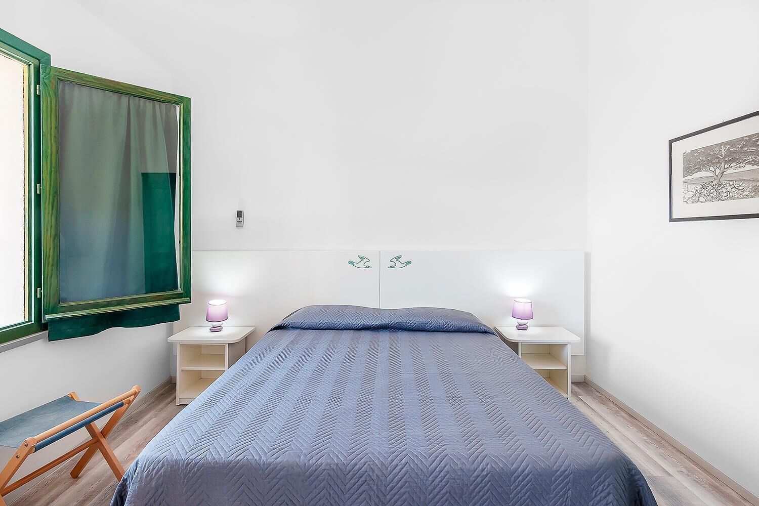 Un dormitorio con una cama, dos mesitas de noche con lámparas, una ventana con cortinas y un cuadro colgado en la pared.