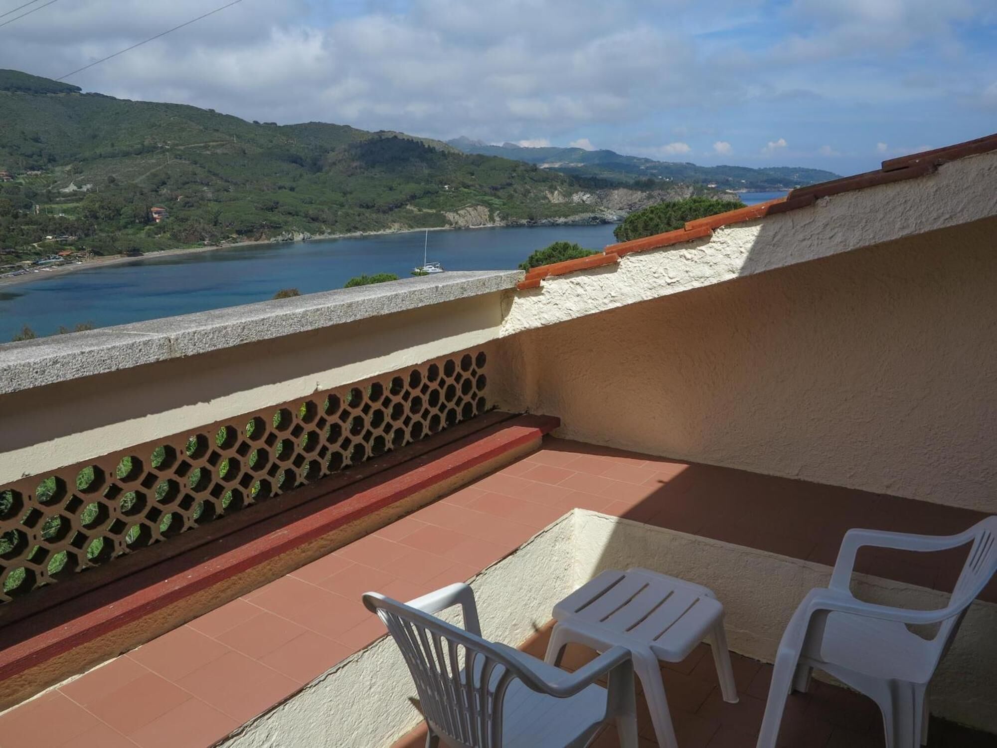 A balcony with two white plastic chairs and a small table, overlooking a body of water and green hills.
