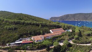 Un hotel costero con varias edificaciones, incluyendo una estructura grande de techo rojo y otras más pequeñas. El hotel está rodeado de vegetación exuberante y da vista a una bahía con barcos.