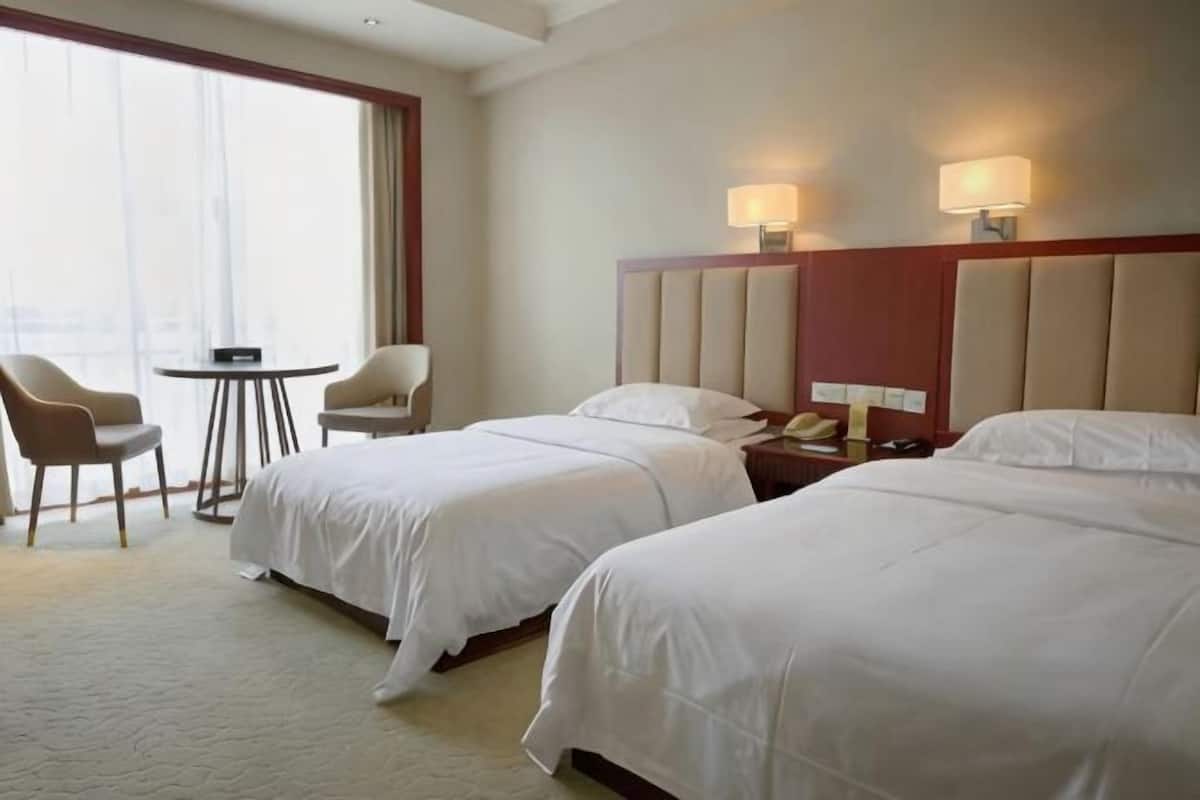 Hotelkamer met twee bedden, een tafeltje, twee stoelen, een nachtkastje met een lamp en een groot raam.
