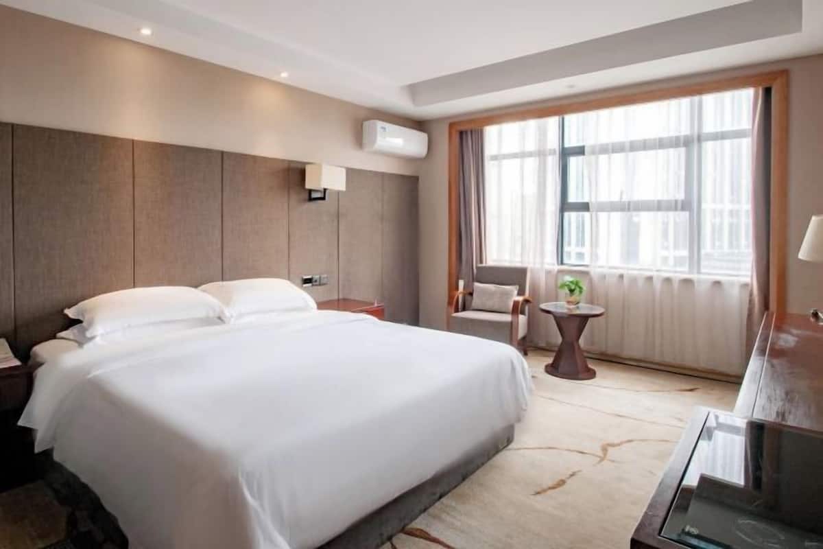 Een hotelkamer met een groot bed, een nachtkastje, een stoel, een kleintje tafeltje met een plant en een raam met gordijnen.
