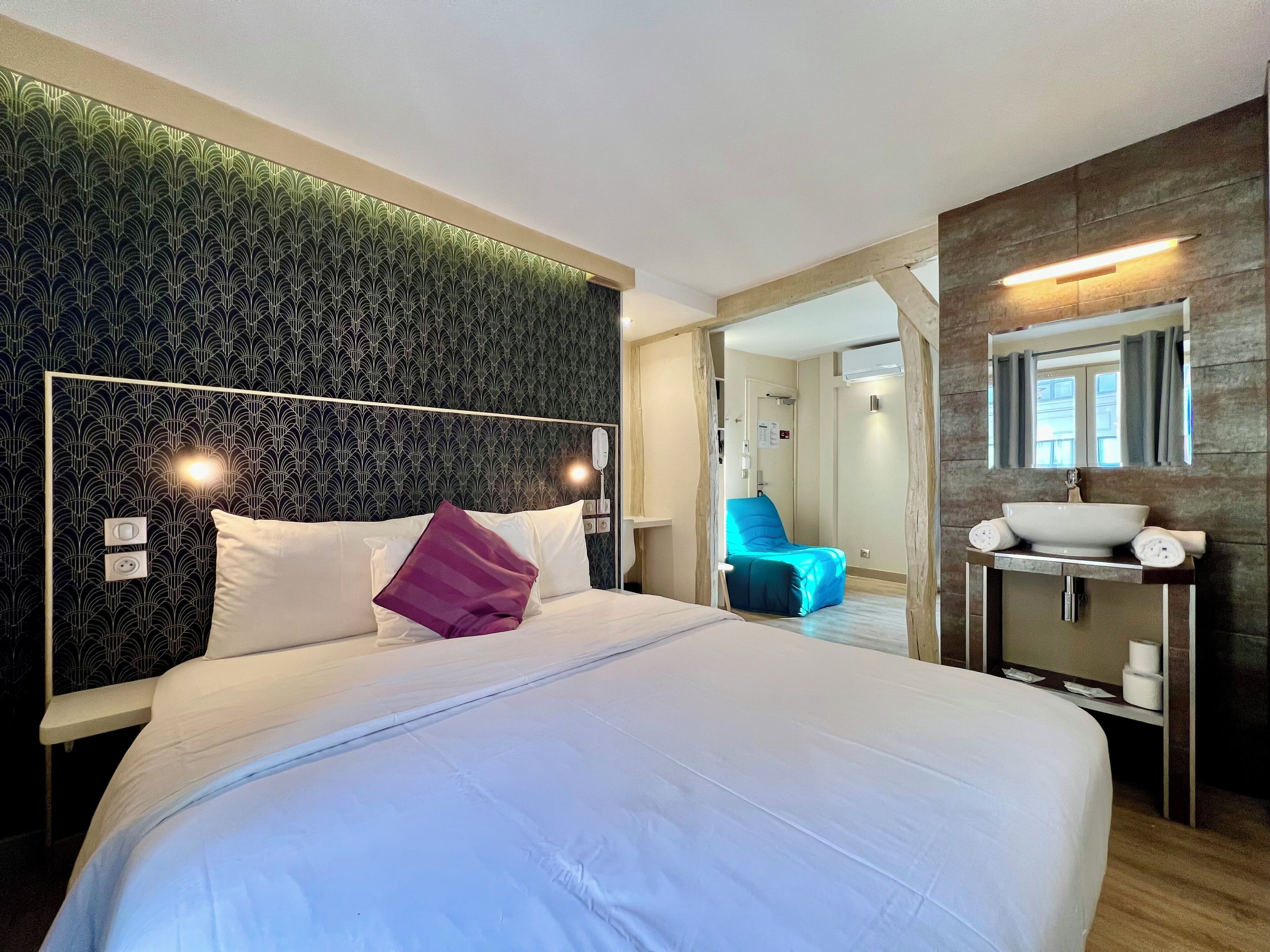 A modern hotel room with a large bed, a blue chair, a vanity with a mirror, and a patterned wall.