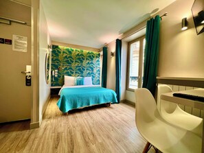 Quarto de hotel com cama, escrivaninha, cadeira, janela com cortinas, parede com papel de parede tropical e porta com placa.