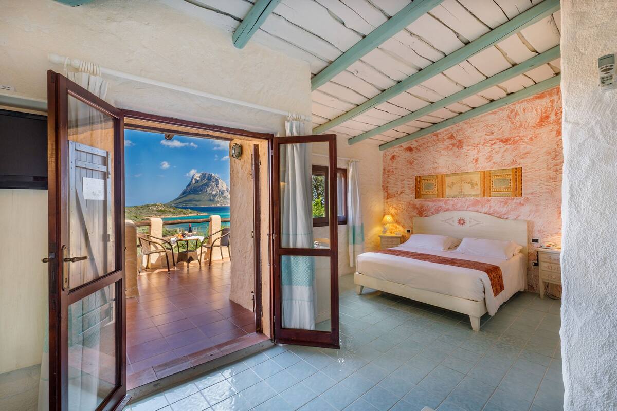 Ein Schlafzimmer mit Bett, Nachttisch und Blick auf eine gebirgige Landschaft durch eine offene Tür.
