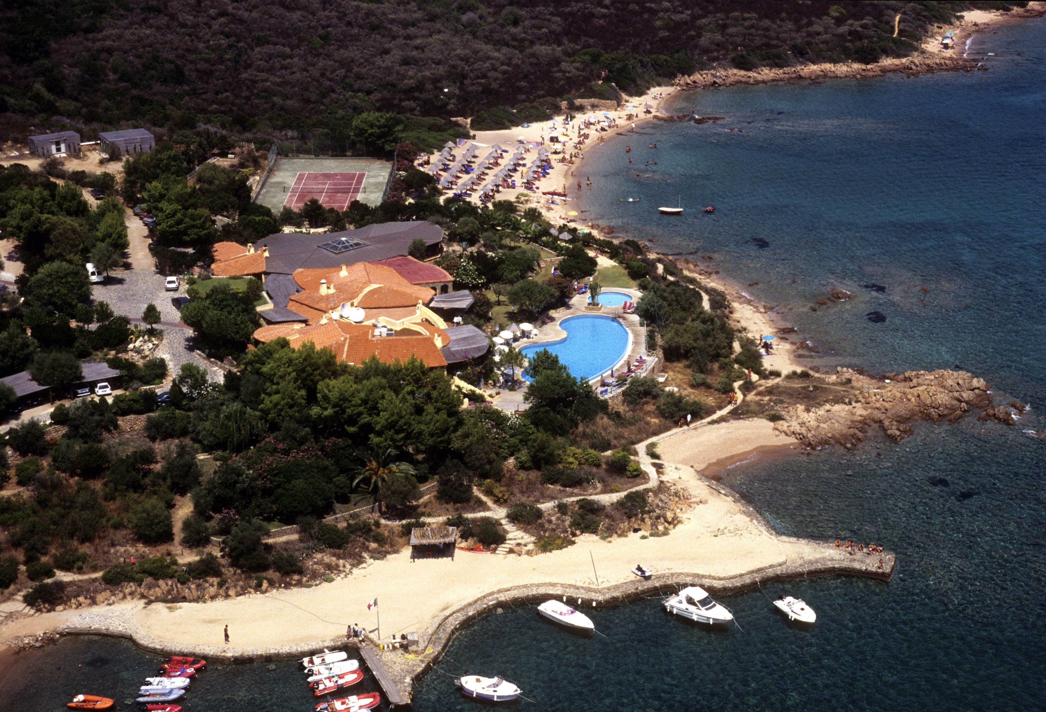 An aerial view of a coastal property with a beach, tennis court, swimming pool, and several boats docked near the shore.
