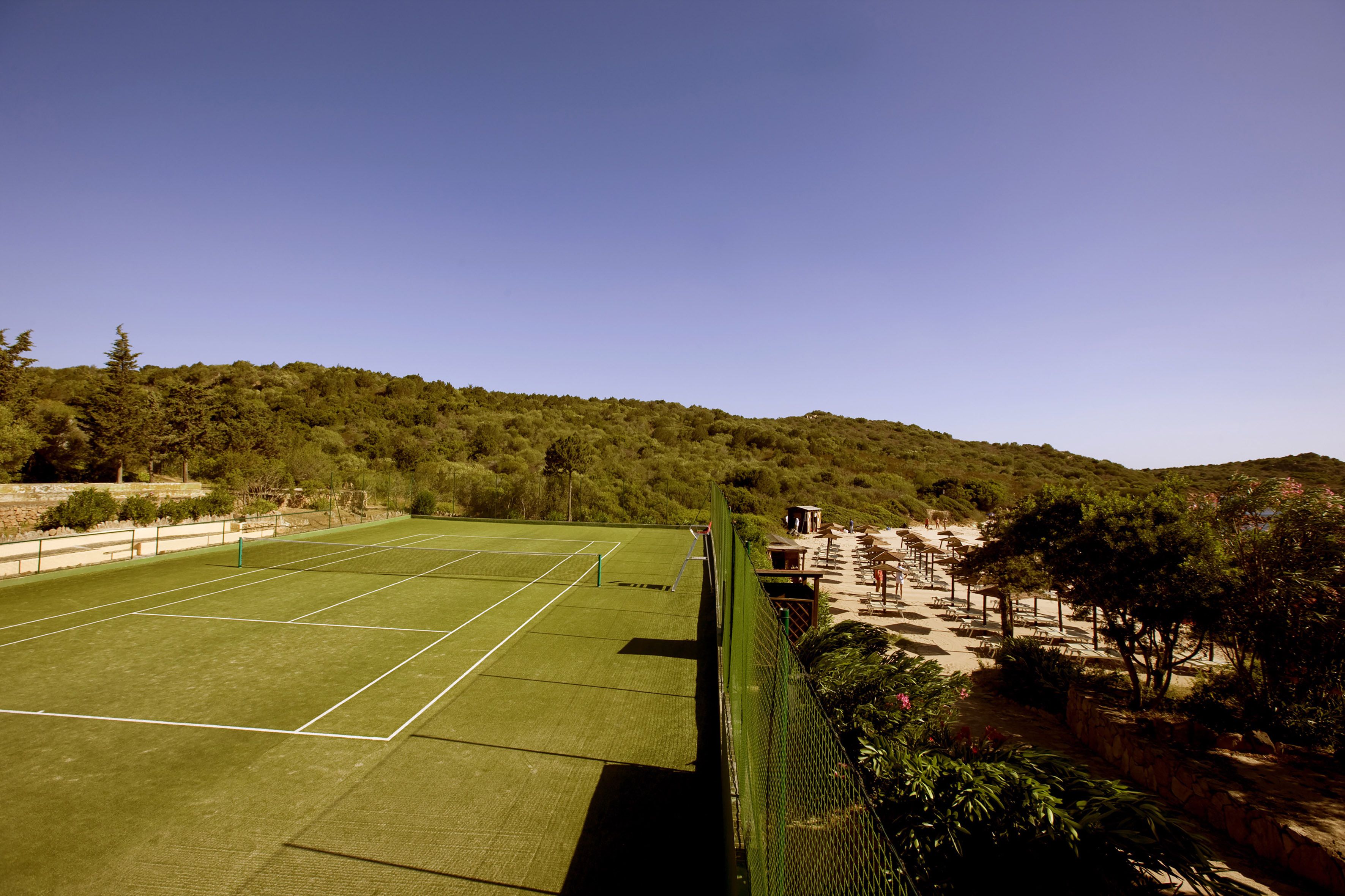 Cancha de tenis con red, rodeada por una valla verde, y un área de descanso con bancos y mesas.