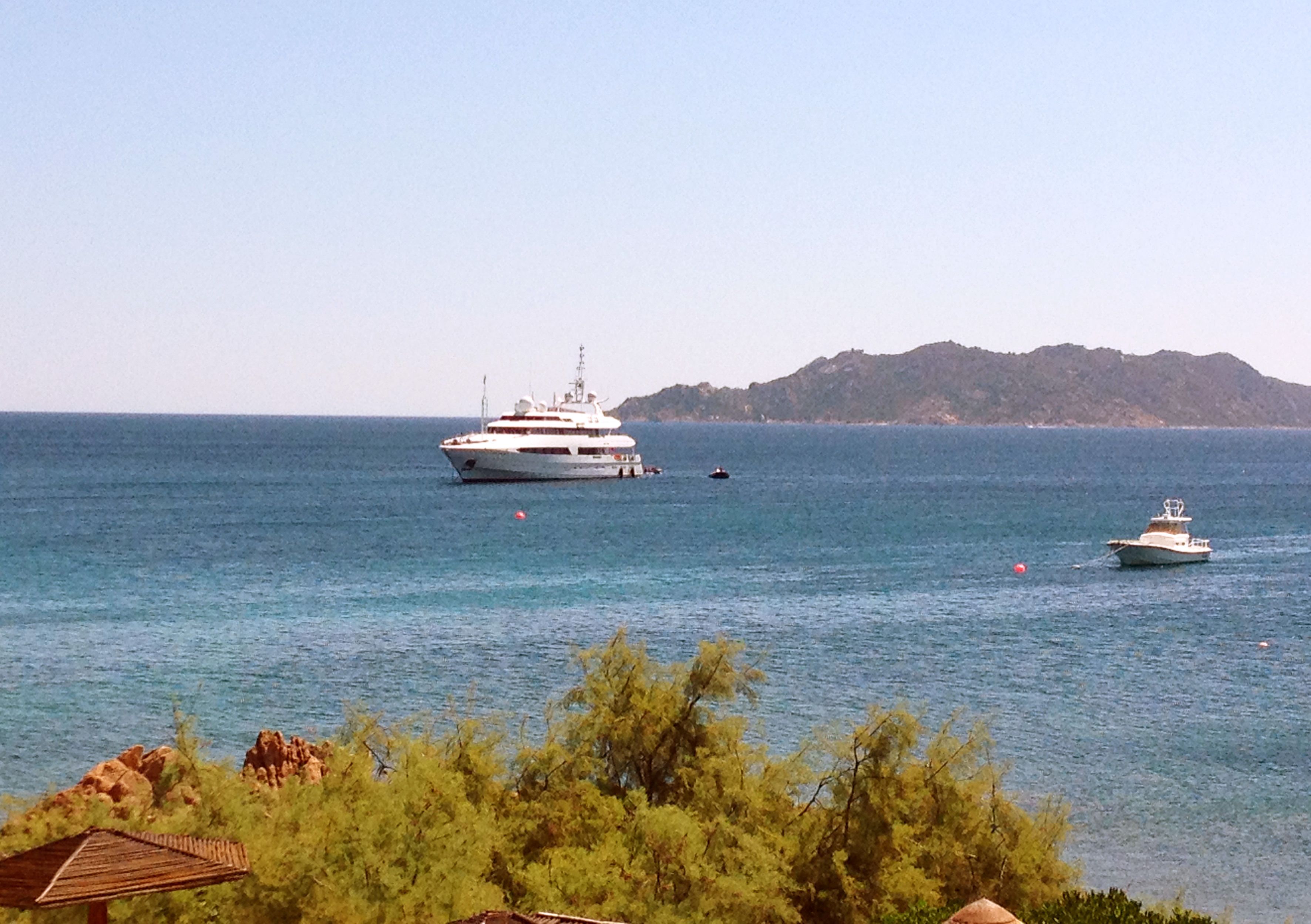 Eine große Yacht auf ruhigem Wasser, in der Nähe ein kleines Boot, eine entfernte Berglandschaft und etwas Grün im Vordergrund.