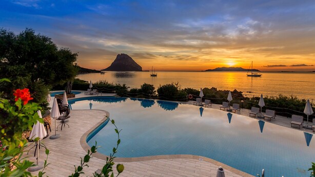 A swimming pool with lounge chairs and umbrellas, surrounded by greenery, with a sunset view over water and a distant mountain.
